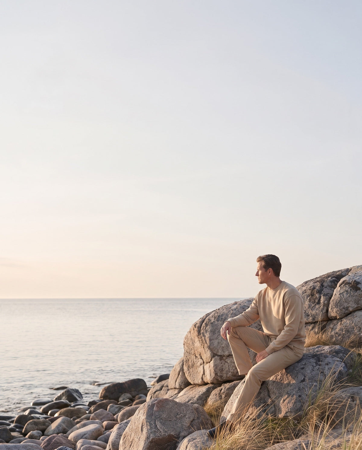 Man in a sand-coloured sweatshirt sitting on coastal rocks in evening light.