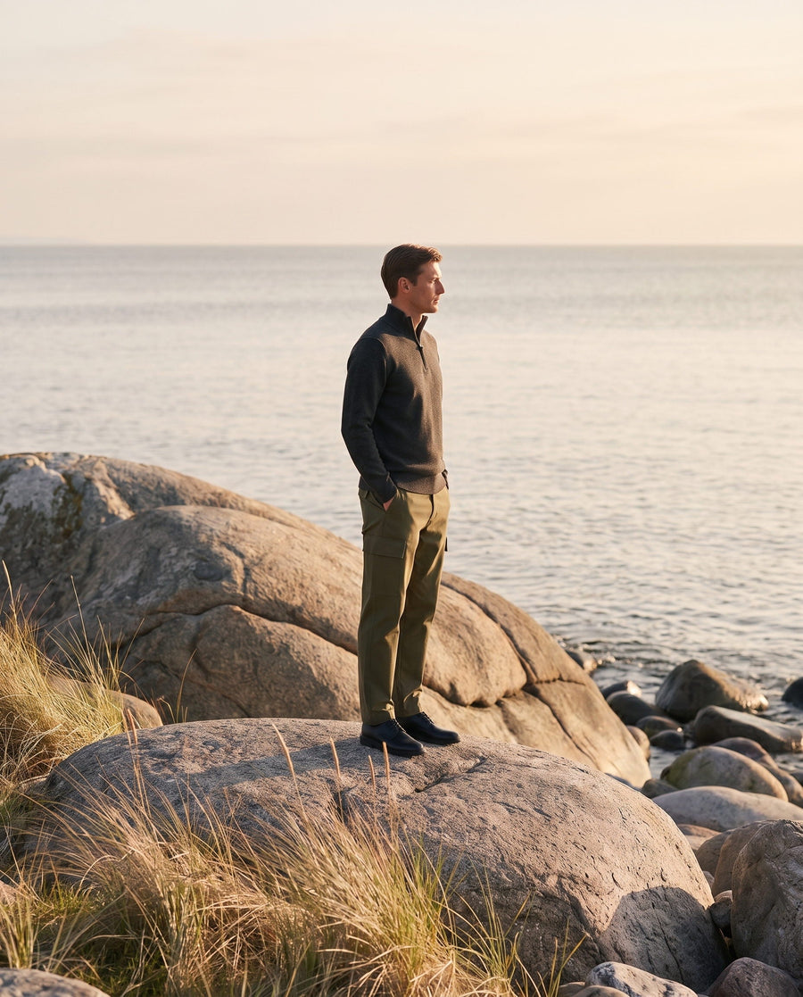 Man in a dark half-zip and olive green trousers standing on coastal rocks by the sea.